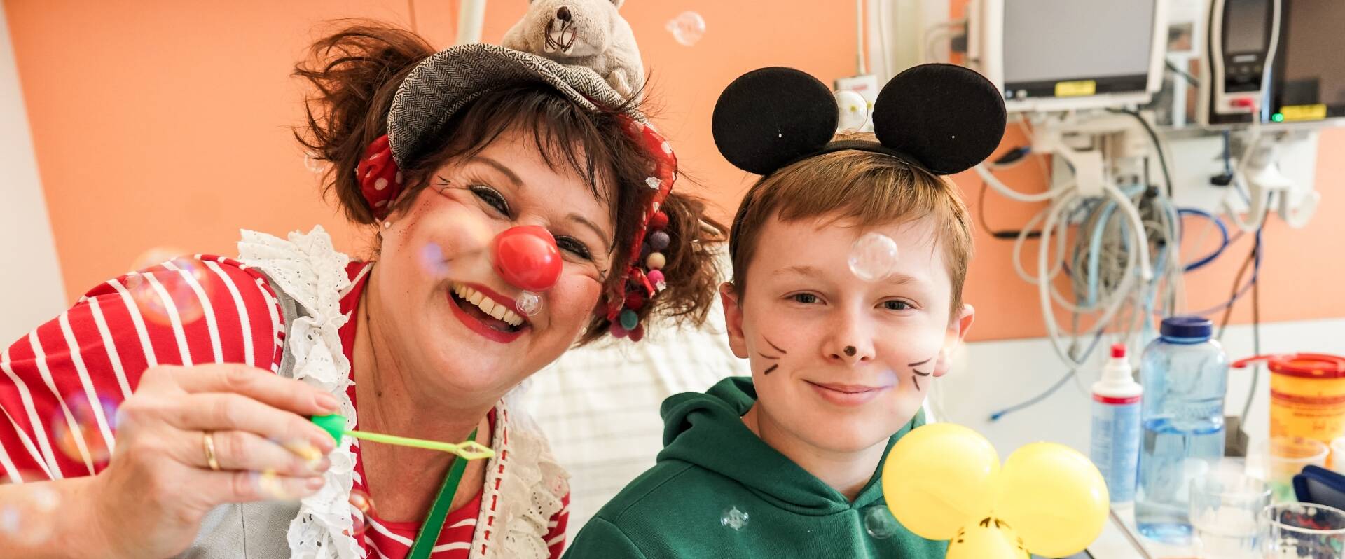Frau als Clown verkleidet, bläst mit einem kleinen Jungen Seifenblasen in die Kamera