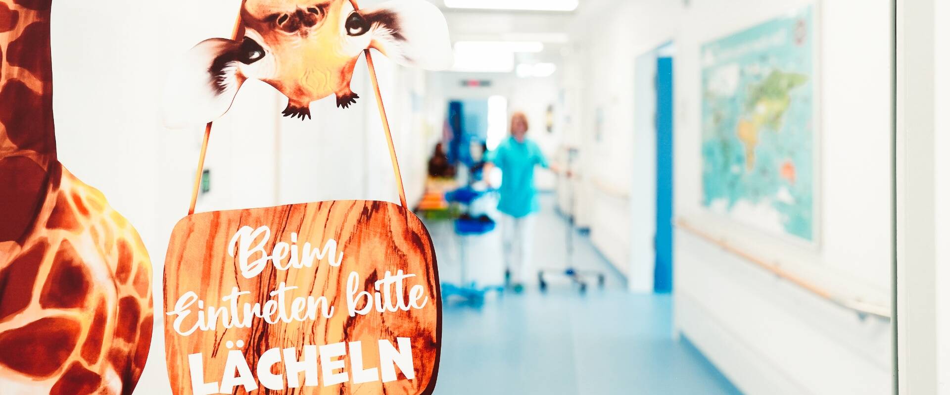 Blick in einen Krankenhausflur, im Vordergrund eine Giraffe, die ein Schild mit der Aufschrift "Beim Eintreten bitte lächeln" trägt