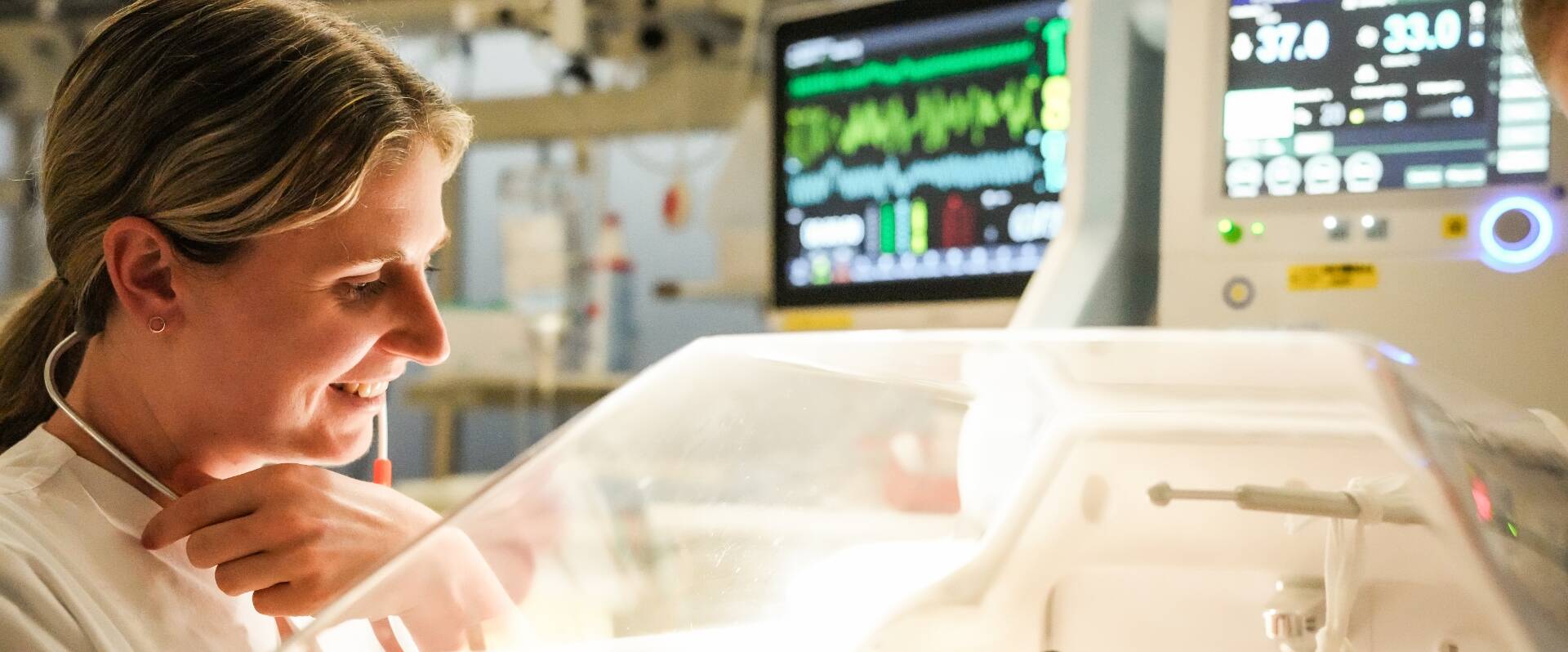 Junge Ärztin in abgedunkeltem Raum lächelt zu Frühchen in Inkubator liegend